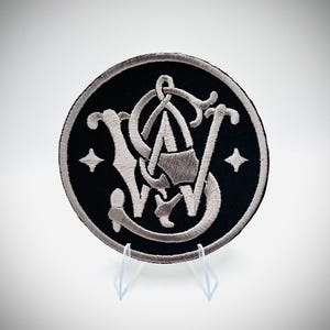 May include: Embroidered patch with a black circular background and a silver-colored logo. The logo features stylized letters "S" and "W" intertwined with a decorative design. Two small star shapes are on either side of the logo.
