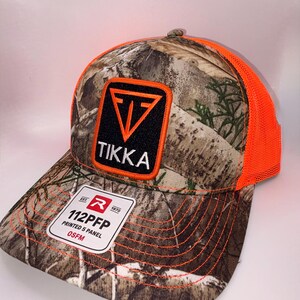 May include: A camouflage print baseball cap with a bright orange mesh back and an orange-trimmed black patch on the front featuring a white Tikka logo. A white rectangular sticker on the brim displays the text 112PFP PRINTED 5 PANEL OSFM.