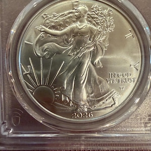 May include: A silver coin featuring Lady Liberty walking towards the sun, with the words "IN GOD WE TRUST" and the year 2026. The coin is encased in a clear protective holder.