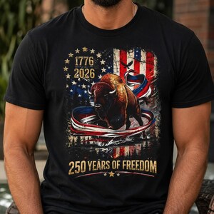 May include: A black t-shirt featuring a graphic of a brown bison centered over a distressed American flag background. The design includes the text 1776, 2026, and 250 YEARS OF FREEDOM, along with a red, white, and blue ribbon swirling around the bison.