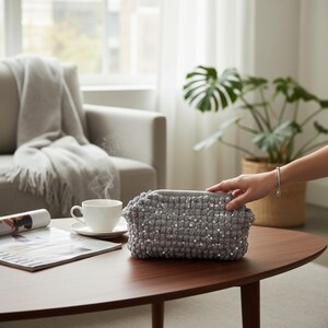 May include: A gray, rectangular, hand-knitted basket with silver sequins is on a dark brown oval coffee table. A white teacup and saucer sit nearby. A person's hand is reaching towards the basket. A gray sofa and a houseplant are in the background.