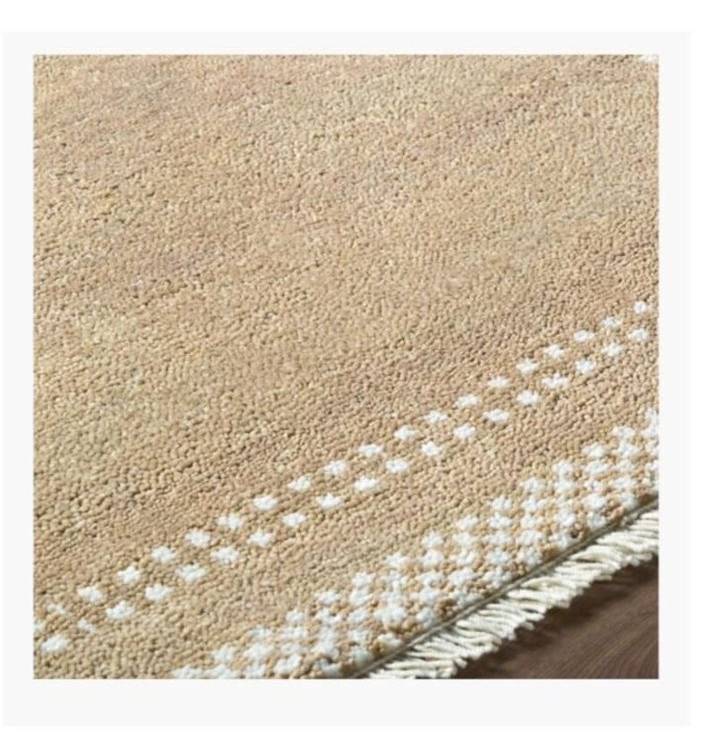 May include: A beige rug with a textured surface and a white dotted border. The rug has a fringe along one edge. The rug is a neutral color and would complement many home decor styles.