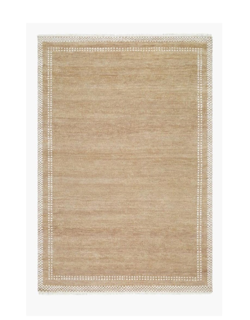 May include: Rectangular beige rug with a white dotted border. The rug has a simple, neutral design, suitable for various interior styles. The texture appears to be woven, adding visual interest. The rug's color is a warm, inviting tone.