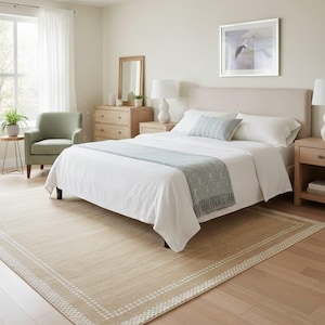 May include: A bedroom scene featuring a bed with white bedding, a light blue throw, and a matching pillow. A beige rug with a white border sits on the light wood floor. The room includes a dresser, side tables, lamps, and a green armchair.
