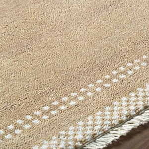 May include: A beige rug with a textured surface and a white dotted border. The rug has a fringe along one edge. The rug is made of a soft material and is suitable for a living room or bedroom.