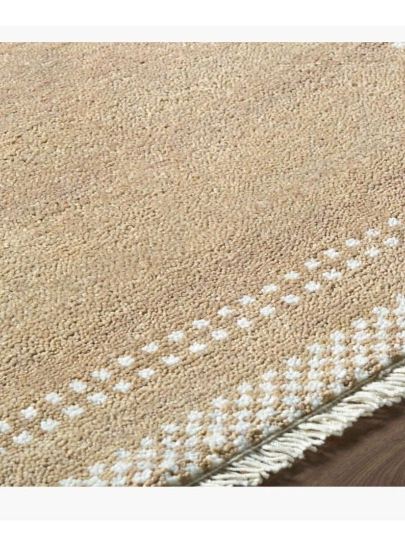 May include: A beige rug with a textured surface and a white dotted border. The rug has a fringe along one edge. The rug is made of a soft material and is suitable for a living room or bedroom.