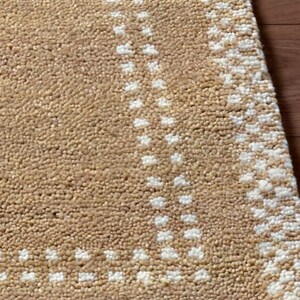 May include: Close-up of a beige rug with a textured surface and a geometric pattern in white. The rug has a fringe along one edge and is partially visible against a wooden surface.