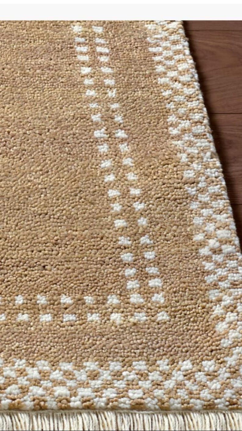 May include: Close-up of a beige rug with a textured surface and a geometric pattern in white. The rug has a fringe along one edge and is partially visible against a wooden surface.