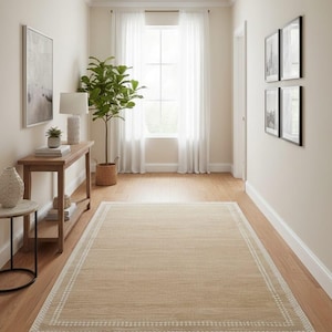 May include: A rectangular beige rug with a white border is centered in a hallway with hardwood floors. The hallway has white walls, a window with sheer white curtains, and a wooden console table with decor.