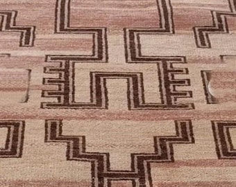 Op de afbeelding: Een rechthoekig tapijt met een herhalend geometrisch patroon in tinten beige, bruin en bruin. Het ontwerp bevat onderling verbonden, trapsgewijze vormen en lijnen, waardoor een getextureerd oppervlak ontstaat. Het tapijt lijkt van geweven materiaal te zijn gemaakt.