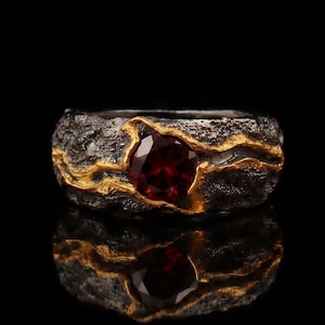 Könnte beinhalten: Ein strukturierter, dunkelgrauer Ring mit goldfarbener Einlage. Der Ring hat einen großen, runden, tiefroten Edelstein. Der Ring wird auf einer reflektierenden schwarzen Oberfläche präsentiert.