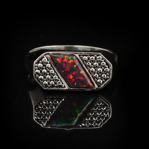 May include: A silver ring featuring a rectangular, red and black opal gemstone. The ring has a geometric design with small, round embellishments. The ring is set against a black background.