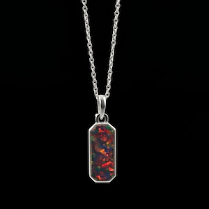 Puede incluir: Un collar de plata con un colgante rectangular de esquinas cortadas. El colgante está incrustado con una piedra oscura e iridiscente que muestra destellos vibrantes de color rojo y naranja. La pieza cuelga de una fina cadena de plata a juego.