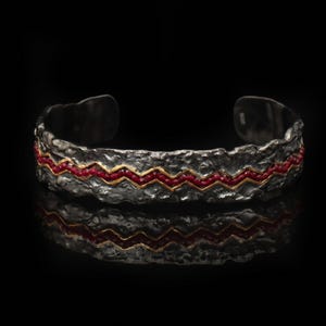 May include: A silver cuff bracelet with a textured surface. The bracelet features a zigzag pattern of red stones and gold accents. The jewelry is displayed on a reflective black surface.