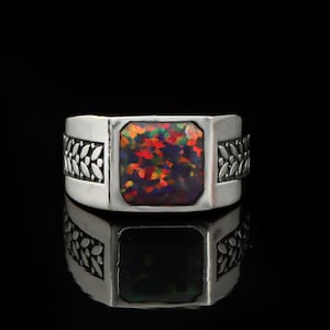 Peut inclure: Une bague en argent avec une pierre d'opale carrée multicolore. La bague présente un motif de feuilles décoratives sur les côtés. L'opale affiche un mélange vibrant de teintes rouges, oranges et vertes, sur fond noir.