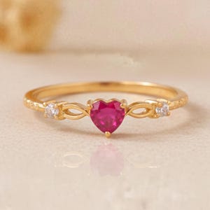 May include: A gold ring featuring a heart-shaped ruby gemstone at the center. The ring has two small, round, clear gemstones on either side of the ruby. The band has a textured design.