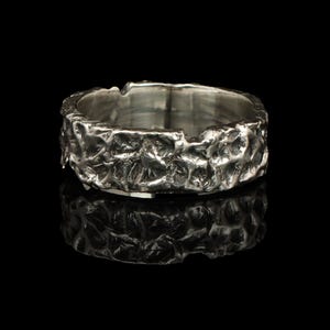 Pode incluir: Um anel de prata texturizado com uma superfície áspera e irregular. O anel tem uma banda larga e um fundo escuro, acentuando a textura metálica. O design do anel sugere uma estética artesanal ou orgânica.