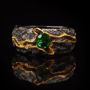 May include: A textured ring with gold detailing and a bright green gemstone. The ring's band has a dark, stone-like texture, with gold-coloured metal outlining the design. The gemstone is round and set in the centre of the ring.