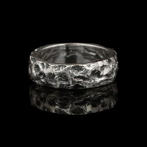 Op de afbeelding: Een zilveren ring met een getextureerd, ongelijkmatig oppervlak. De ring heeft een bandachtig ontwerp met een ruwe, organische textuur die licht reflecteert. De ring staat tegen een zwarte achtergrond, met zijn weerspiegeling eronder zichtbaar.