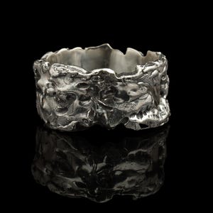 Puede incluir: Un anillo plateado texturizado con una superficie irregular. El anillo presenta un diseño rugoso y esculpido, que captura la luz y crea sombras. Se muestra sobre un fondo negro, resaltando su brillo metálico. El anillo mide aproximadamente 1,5 cm de ancho.
