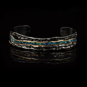 Op de afbeelding: Een getextureerde zilveren manchetarmband met een gouden band en een rij blauwgroene opalen. De armband heeft een donkere, gehamerde uitstraling en wordt gepresenteerd op een zwarte ondergrond.