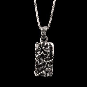Puede incluir: Collar con colgante plateado con un colgante rectangular texturizado. El colgante cuelga de una delicada cadena sobre un fondo negro. El colgante tiene una superficie rugosa e irregular.