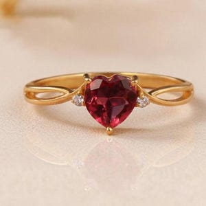 May include: A gold ring featuring a heart-shaped, faceted, deep red gemstone. The ring has two small, round, clear gemstones on either side of the heart. The band has an open, looping design. A romantic piece of jewelry.