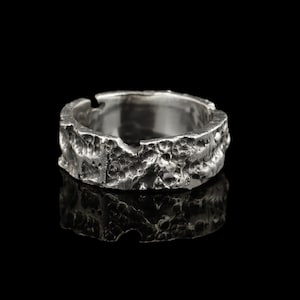 May include: A silver ring with a textured, rocky design. The ring has a rough, uneven surface with a small cut-out. The ring is shown against a black background, emphasizing its metallic shine and unique texture.