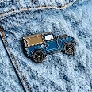 Könnte beinhalten: Eine blaue und braune Emaille-Anstecknadel mit einem Vintage Land Rover Defender. Die Anstecknadel befindet sich auf einem blauen Jeans-Hintergrund.