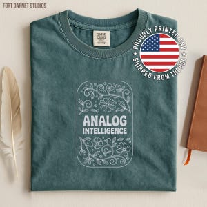 May include: A teal Comfort Colors t-shirt with a white floral design and the words "ANALOG INTELLIGENCE." The shirt has a crew neck and a graphic that says "PROUDLY PRINTED AND SHIPPED FROM THE USA."
