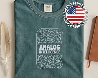 May include: A teal Comfort Colors t-shirt with a white floral design and the words "ANALOG INTELLIGENCE." The shirt has a crew neck and a graphic that says "PROUDLY PRINTED AND SHIPPED FROM THE USA."