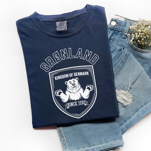 May include: Navy blue t-shirt with a white graphic of a bear inside a shield, the text "GRØNLAND KINGDOM OF DENMARK SINCE 1721". The shirt is paired with denim shorts and a small bouquet of white flowers.