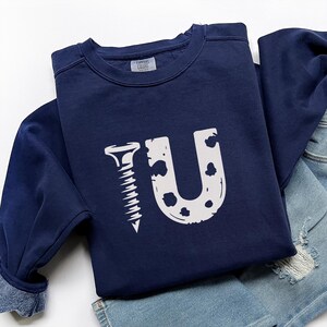 May include: Navy blue sweatshirt with a white graphic that spells out "Screw U". The design features a screw replacing the letter "I" and a dotted "U". The sweatshirt is folded on top of a pair of denim shorts.