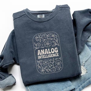 May include: A navy blue sweatshirt with the words "ANALOG INTELLIGENCE" in a floral design. The sweatshirt is folded on top of a pair of blue jeans. The brand "Comfort Colors" is printed on the upper back of the sweatshirt.