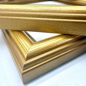 May include: Close-up of two gold-colored picture frames. The frames are made of wood and have a textured, metallic finish. The corners are mitered, and the frames are stacked at an angle, showing the details of the molding.