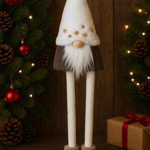 Op de afbeelding: Een decoratieve kerstkabouter met een witte hoed versierd met houten knopen en een lange witte baard. De kabouter heeft lange, gestreepte benen en draagt grijze laarzen. De achtergrond omvat een kerstboom met rode ornamenten en een ingepakt cadeau.