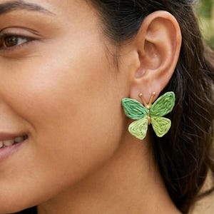 May include: A pair of butterfly-shaped stud earrings featuring textured green thread wings and a gold-toned metal center body. The wings display a gradient of light and dark green shades, with a small gold antenna detail at the top of the butterfly.
