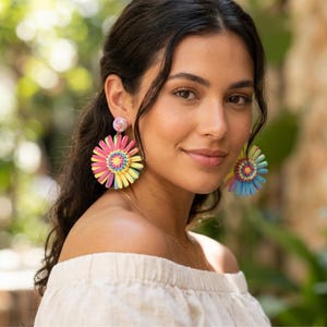May include: A pair of large, circular statement earrings featuring a vibrant, multi-colored floral design. The earrings are crafted with layered, radiating petals in shades of bright pink, yellow, and blue, centered with a small, textured stud.