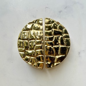 Op de afbeelding: Twee halve cirkel kastgrepen in een heldere gouden afwerking. De grepen hebben een textuur, baksteenachtig patroon over hun oppervlak. De hardware wordt getoond tegen een witte marmeren achtergrond.