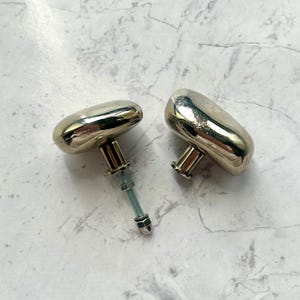 May include: Two metallic cabinet knobs with a smooth, rounded, pebble-like shape and a polished gold-tone finish. One knob features a threaded metal bolt extending from the base, complete with a small nut and washer attached to the hardware.