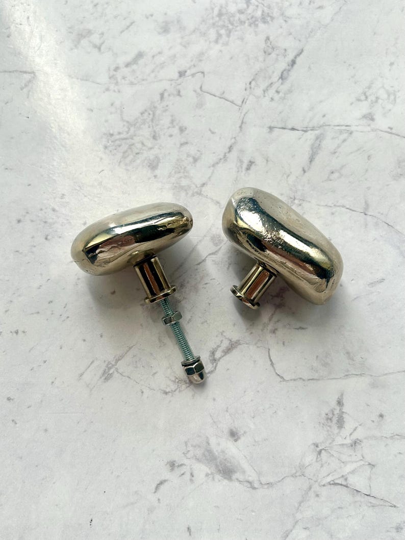 May include: Two metallic cabinet knobs with a smooth, rounded, pebble-like shape and a polished gold-tone finish. One knob features a threaded metal bolt extending from the base, complete with a small nut and washer attached to the hardware.