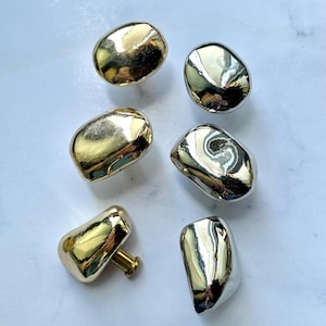 May include: A collection of six cabinet knobs with irregular, organic pebble-like shapes. Three knobs feature a polished gold-tone finish, while the other three have a reflective silver-tone finish. Each knob includes a short cylindrical mounting post.