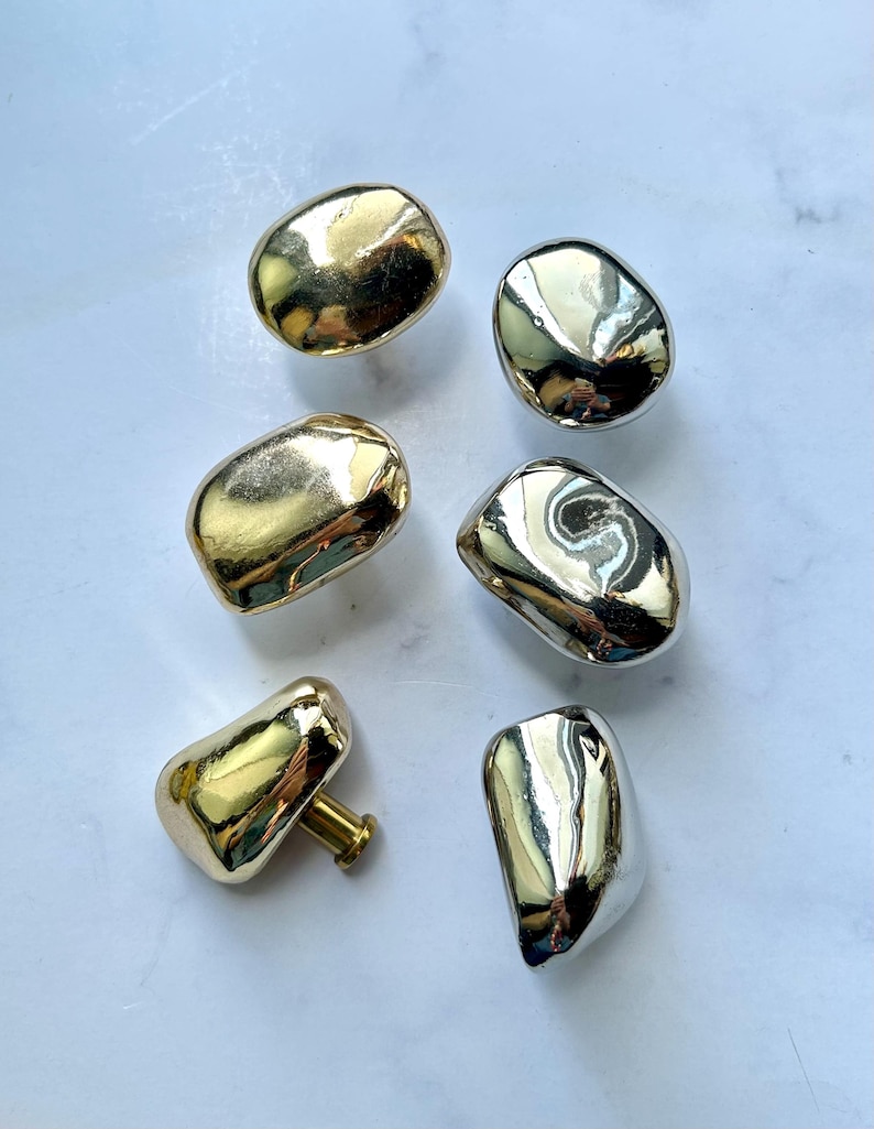 May include: A collection of six cabinet knobs with irregular, organic pebble-like shapes. Three knobs feature a polished gold-tone finish, while the other three have a reflective silver-tone finish. Each knob includes a short cylindrical mounting post.