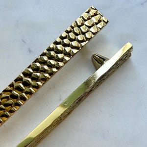 May include: Close-up of two gold-coloured cabinet pulls. One pull has a textured, honeycomb-like pattern, while the other has a more linear, rough texture. The pulls are set against a white and grey marble background.