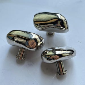 May include: Three cabinet knobs with a polished silver-tone finish and an irregular, organic, pebble-like shape. Each knob features a smooth, reflective surface and a cylindrical base for mounting, showcasing a metallic luster and rounded edges.