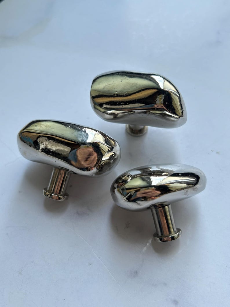 May include: Three cabinet knobs with a polished silver-tone finish and an irregular, organic, pebble-like shape. Each knob features a smooth, reflective surface and a cylindrical base for mounting, showcasing a metallic luster and rounded edges.