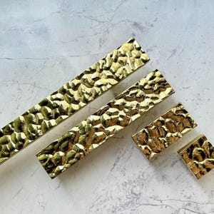 May include: A collection of gold-colored cabinet hardware with a hammered texture. The set includes long rectangular pulls and smaller square and rectangular knobs. The hardware is arranged on a light gray and white marble-like surface.