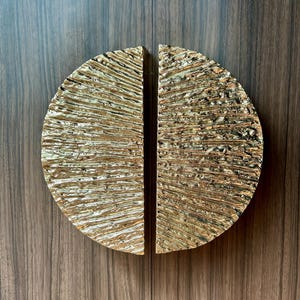 Può includere: Due maniglie dorate per armadi a semicerchio con finitura martellata e strutturata. Le maniglie sono montate su un'anta di un armadio in legno scuro. Le maniglie sono progettate per creare un cerchio completo quando sono chiuse.