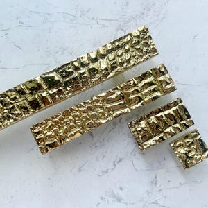 Puede incluir: Un conjunto de herrajes dorados para armarios con un patrón texturizado similar a la piel de cocodrilo. El conjunto incluye tiradores rectangulares largos y pomos cuadrados más pequeños, todos con un acabado brillante. Los herrajes están dispuestos sobre una superficie clara.