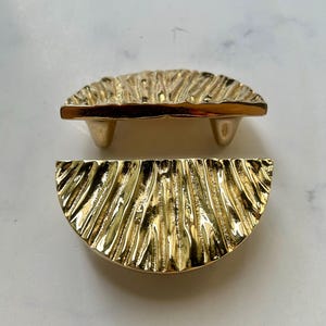 May include: Two gold-colored, half-circle cabinet pulls with a textured, ridged surface. One pull is upright with three small feet, and the other is lying flat. The pulls are made of a shiny, metallic material.